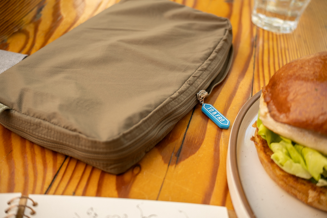 A close-up of a TO&FRO organizer protecting the TRAVELER'S notebook beside an iced tea and plate with a chicken burger and fries.