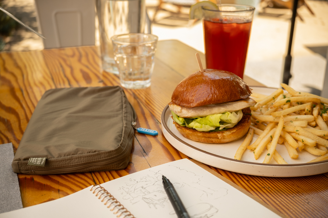 A wooden restaurant table with a TO&FRO organizer protecting the TRAVELER'S notebook beside an iced tea and plate with a chicken burger and fries.