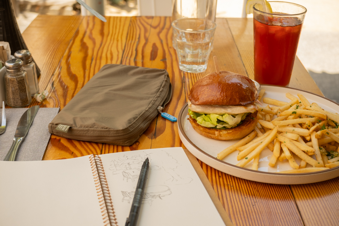 A wooden restaurant table with a TO&FRO organizer protecting the TRAVELER'S notebook beside an iced tea and plate with a chicken burger and fries.