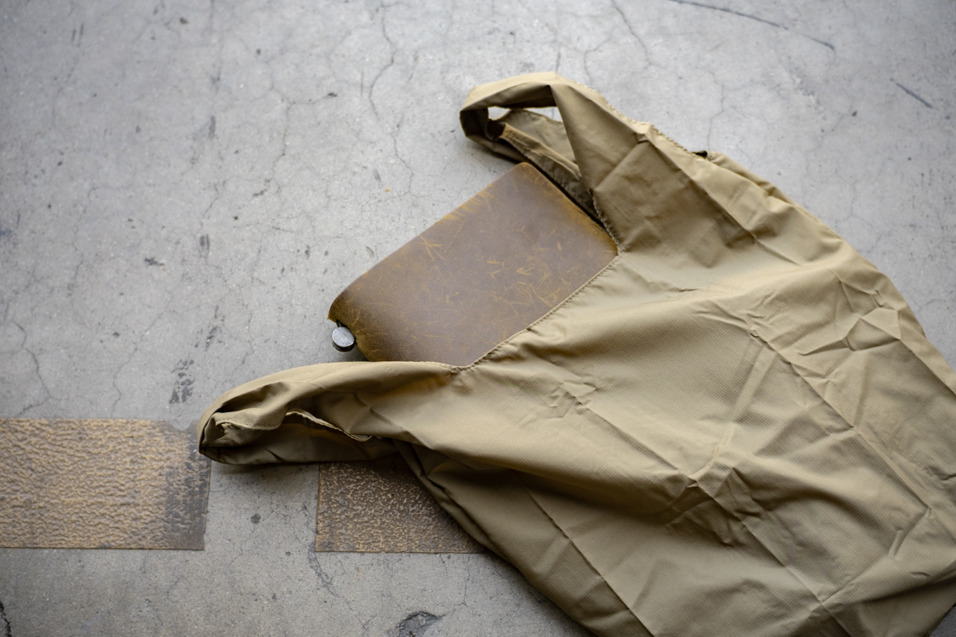 The TO&FRO compact bag in olive stuffed with a TRAVELER'S notebook on a concrete surface.