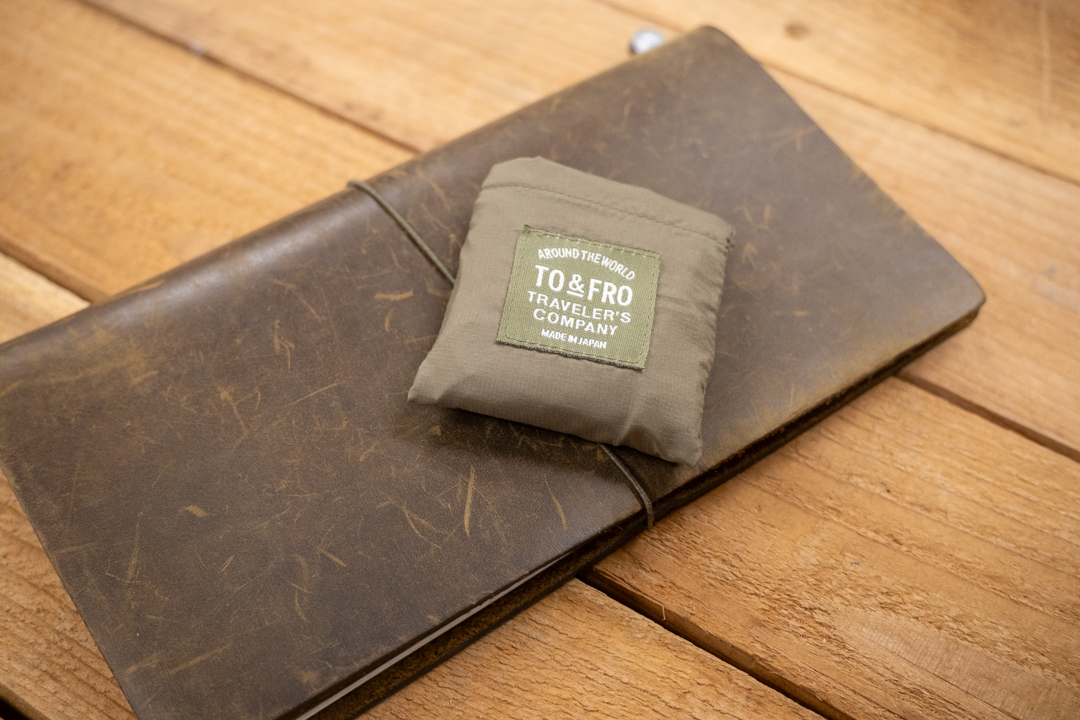 TO&FRO compact bag packed up into a small pocket on top of an Olive TRAVELER'S notebook Regular Size on a wooden table.