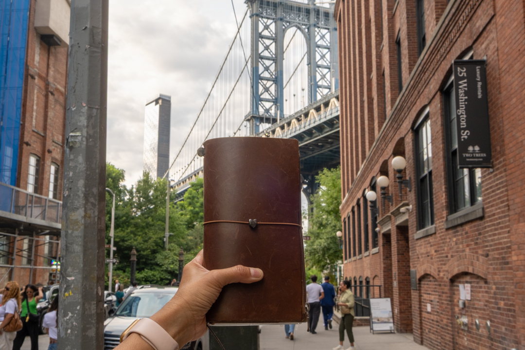 A person holding a TRAVELER'S notebook in front of the view of the Manhattan Bridge from Dumbo, Brooklyn.