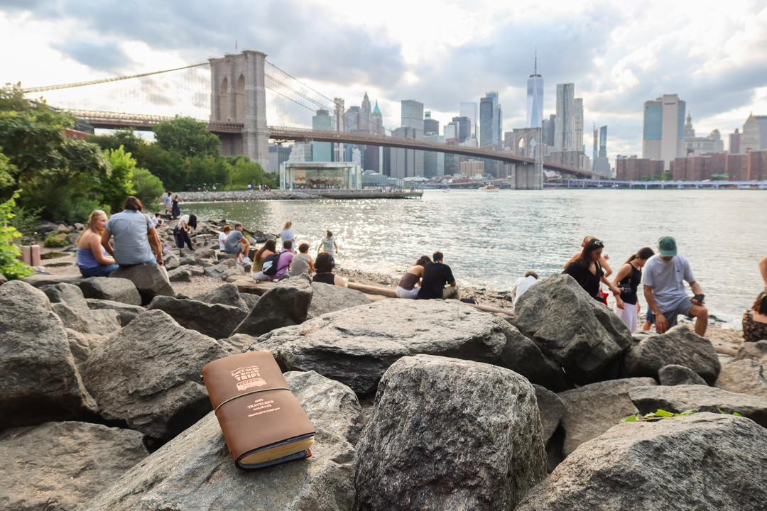 A view of the Brooklyn Bridge from Dumbo, Brooklyn, with a TRAVELER'S notebook resting on a large boulder in the foreground.