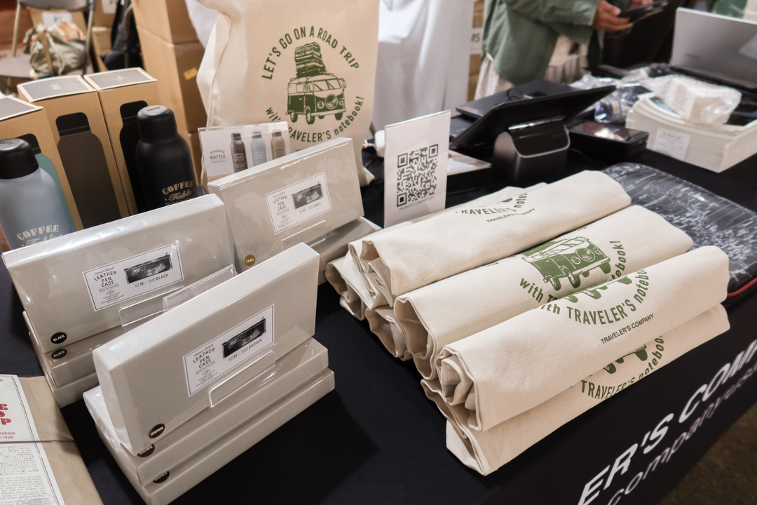 A stack of TRC leather pen cases beside the pile of rolled-up tote bags.